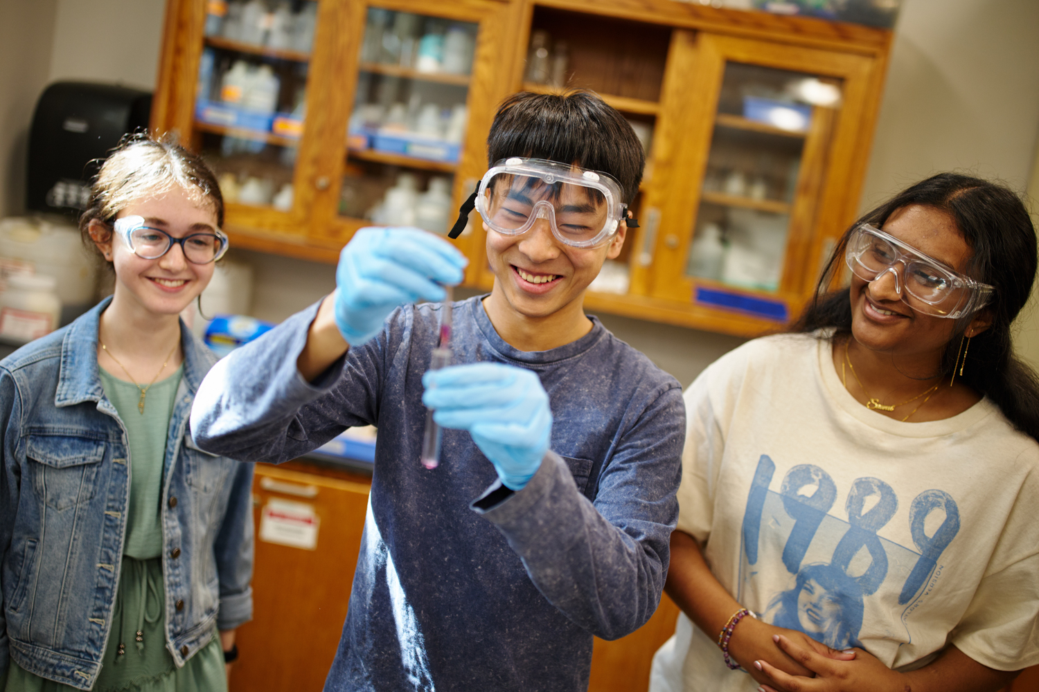 Three students in science goggles and gloves in a lab, with one student holding up a test tube