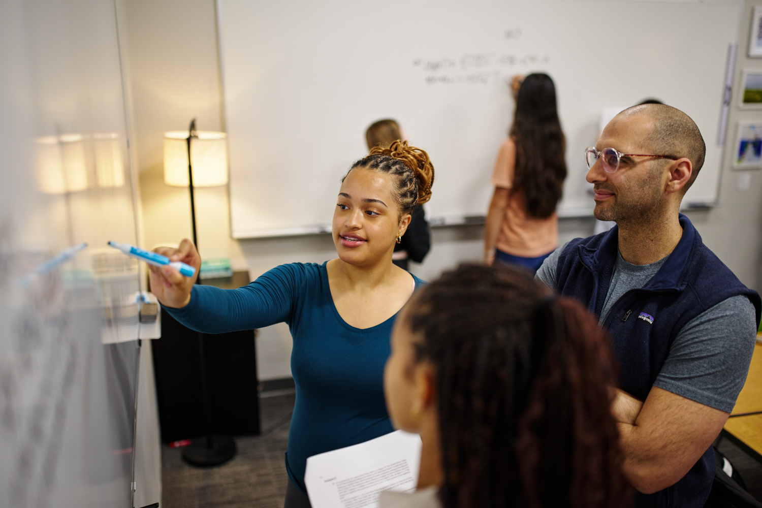 A student and a teacher doing math at a white board with students in the background