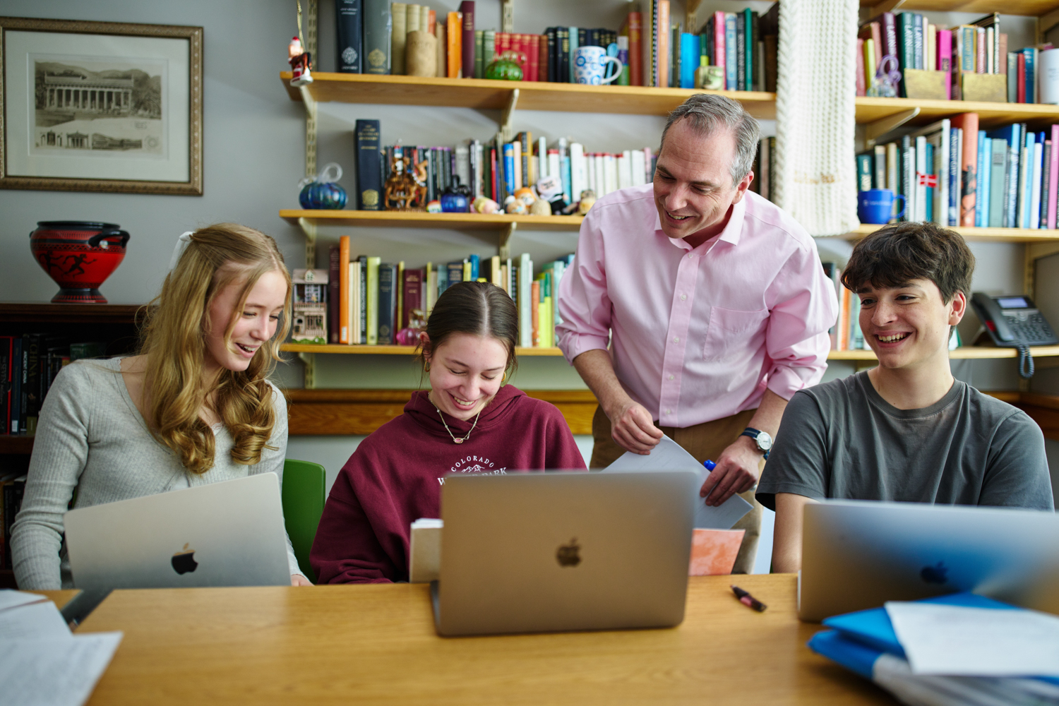 A group of three students laughing and a teacher looking over their shoulders at a book