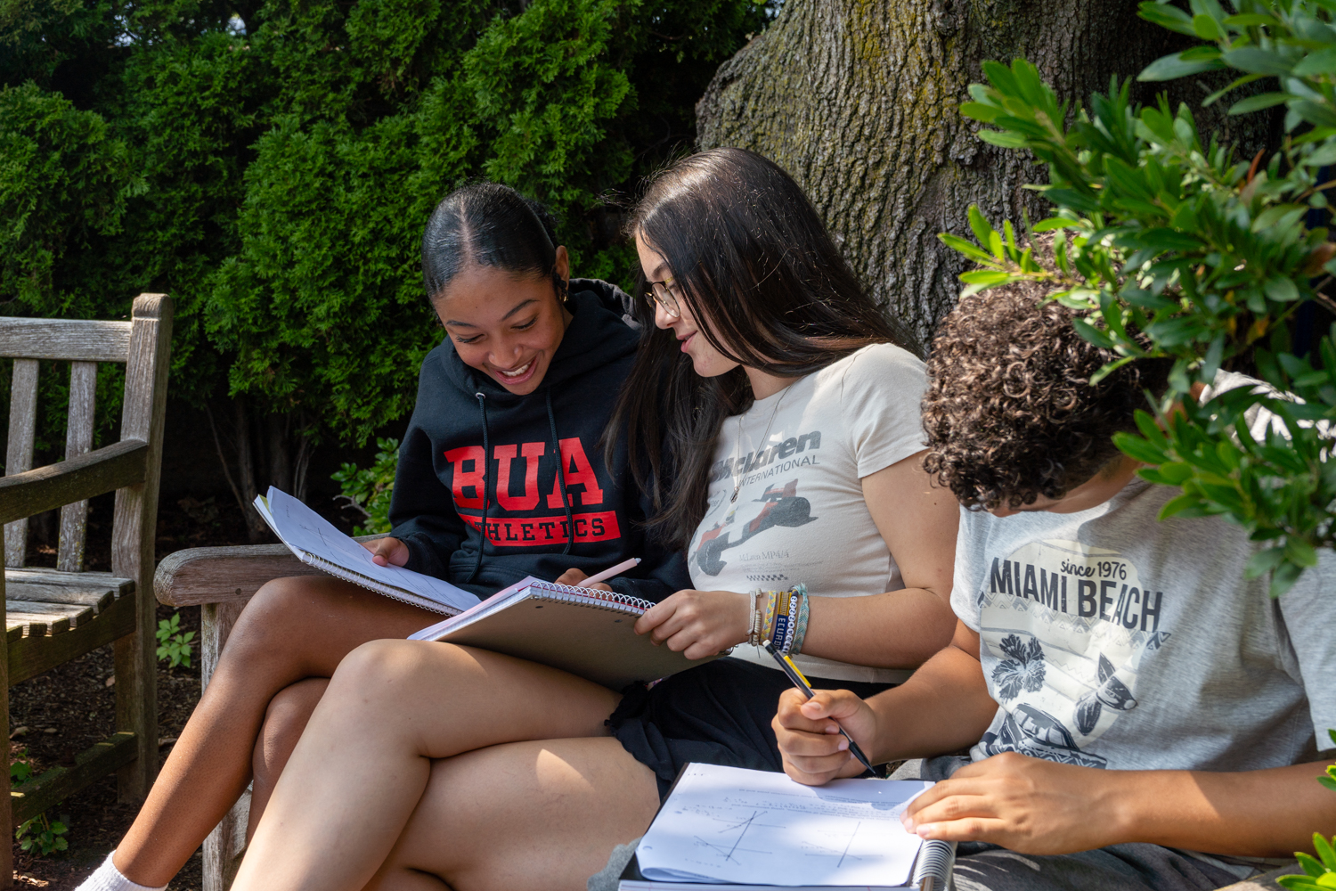 Students working on a bench outside, one student laughing and pointing at the other student's book as they work together